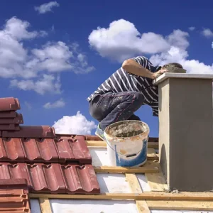 A worker performing chimney repairs on a tiled roof under a blue sky, demonstrating the importance of chimney and roof maintenance.